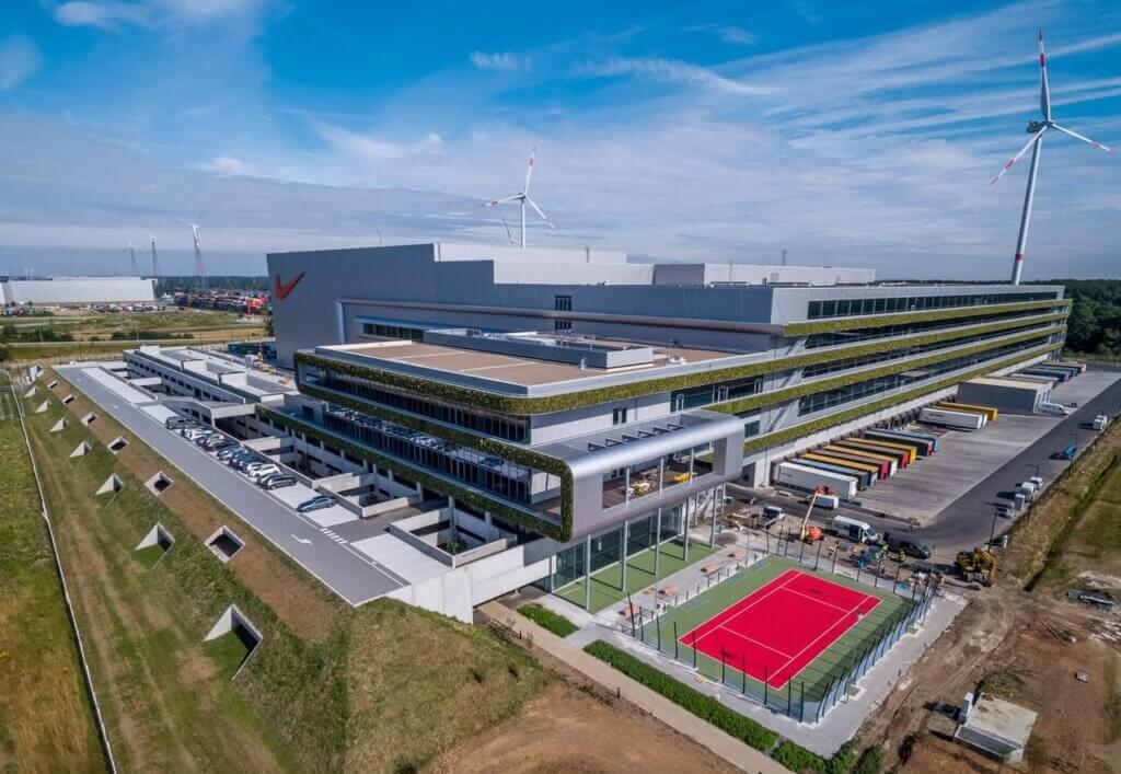 Image of the Nike European Logistics Center. The focus on the picture is sustainability: there is a lot of green space shownon the building, as well as solar panels and wind energy. The logistics service provider trucks are shown on the right side of the picture. The warehouse has very modern architecture.